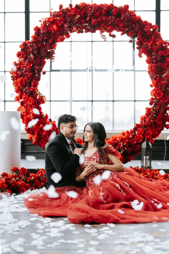 Sonali and Gagan Sitting Under Red Floral Arch with Falling Petals